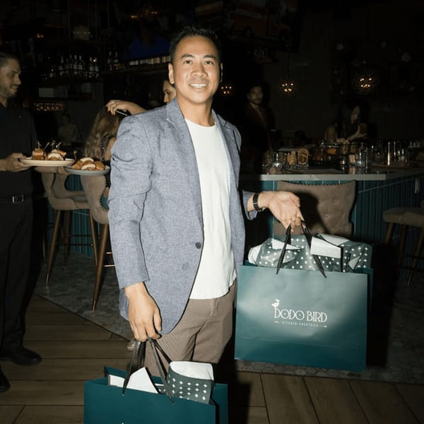 Man smiling, holding Dodo Bird Kitchen and Cocktails branded bags, with a server and appetizers in the background.
