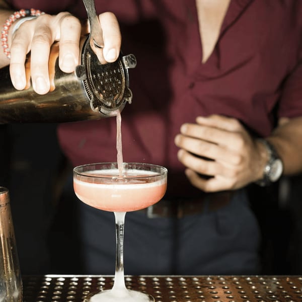 A bartender in a maroon shirt pours a pink cocktail from a shaker into a coupe glass.
