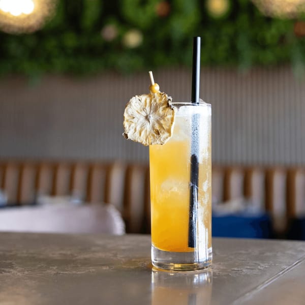 A tall glass of orange cocktail with ice, a black straw, and a dried pineapple garnish.