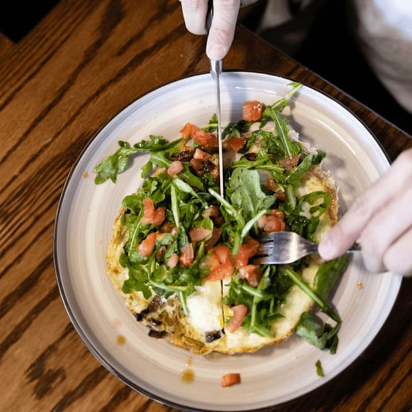 Hands cutting into a Dodo Bird Kitchen omelet topped with fresh arugula and diced tomatoes.