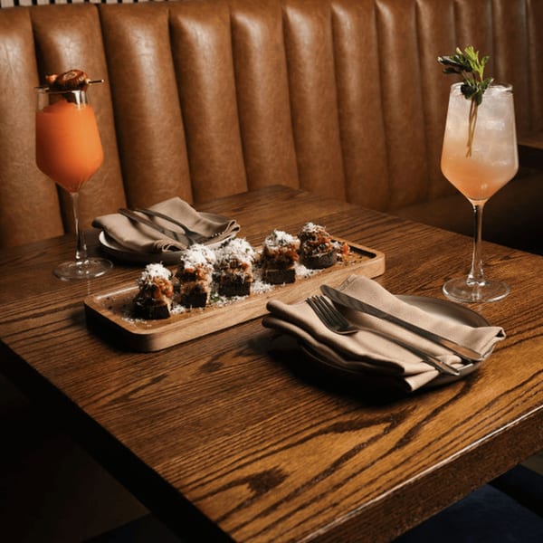 Two cocktails and a wooden board with six appetizers on a dark wooden table with a brown leather booth.