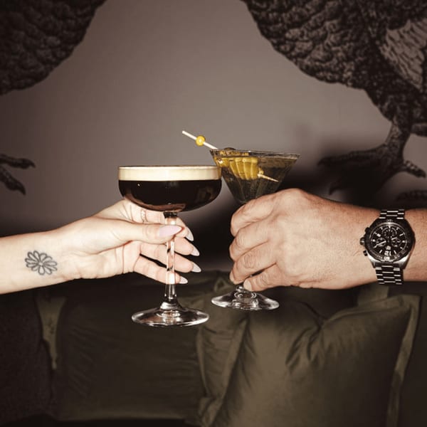 Two hands clinking an espresso martini and a regular martini with olives, against a dark background.