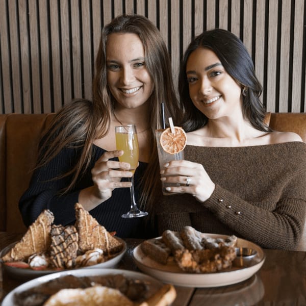 Two smiling women at Dodo Bird Kitchen and Cocktails, holding cocktails with dessert plates in front.