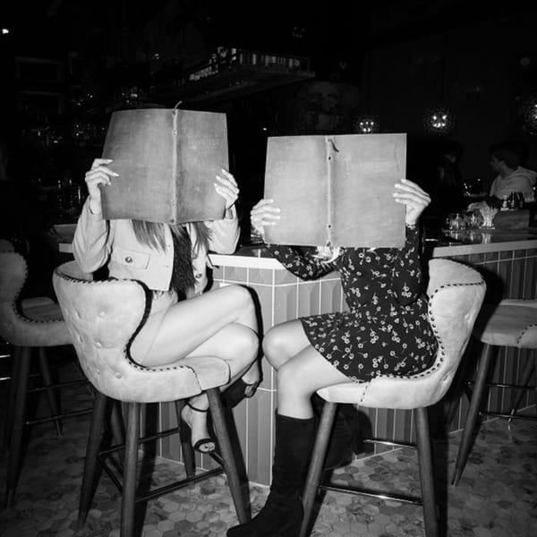Two women at Dodo Bird Kitchen and Cocktails bar, hiding faces behind large menus.