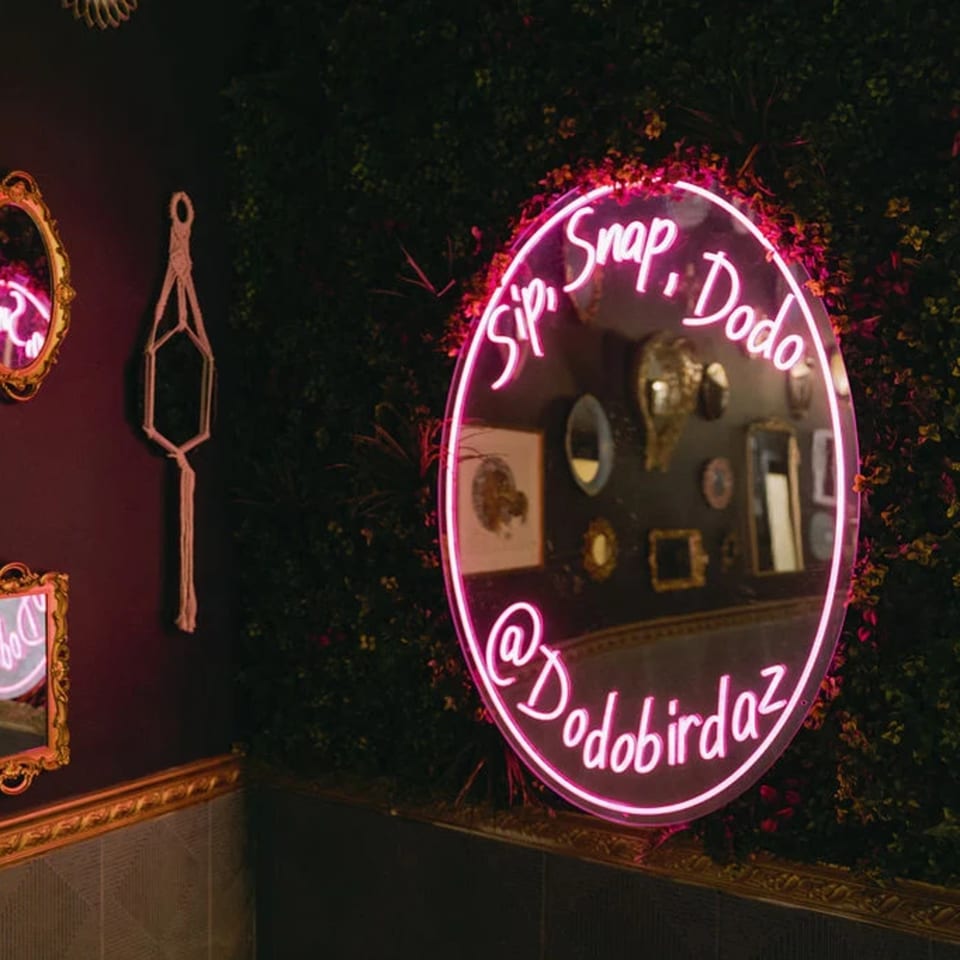 Pink neon sign on a green plant wall, reflecting in a mirror, with "Sip, Snap, Dodo @Dodobirdaz".