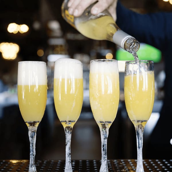 A bartender pours sparkling wine into a mimosa glass, completing a row of four drinks at Dodo Bird Kitchen.