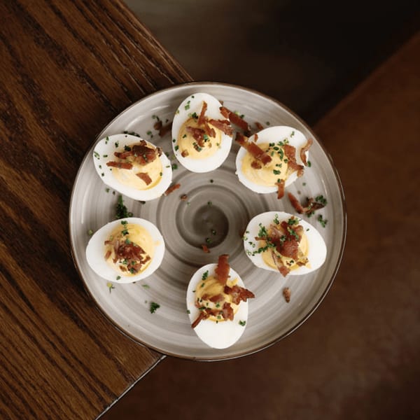 Six deviled eggs with creamy yellow filling, topped with bacon and chives, on a gray plate.