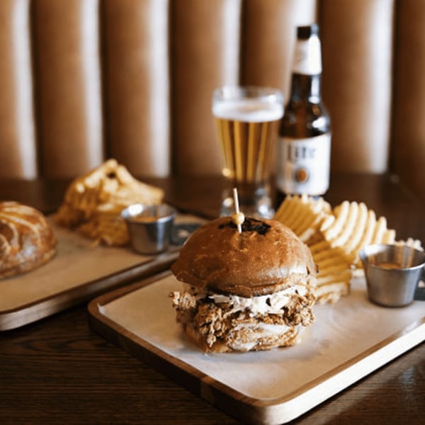A Dodo Bird Kitchen pulled pork sandwich with waffle fries, beer, and a second dish on a wooden table.