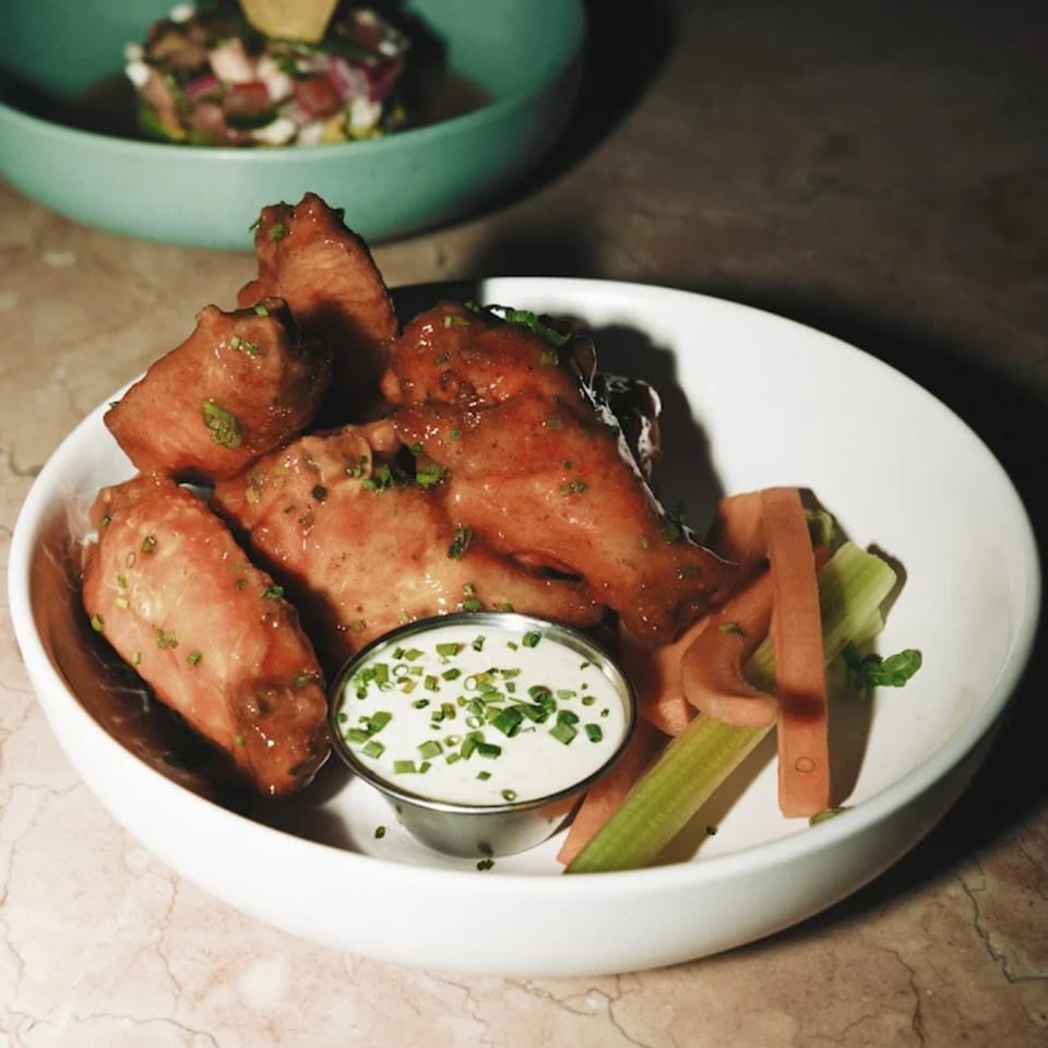 A white bowl of Dodo Bird Kitchen and Cocktails' chicken wings with dipping sauce and vegetable sticks.