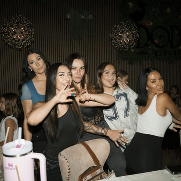 Five women posing playfully at Dodo Bird Kitchen and Cocktails, with decorative lighting and signage in the background.
