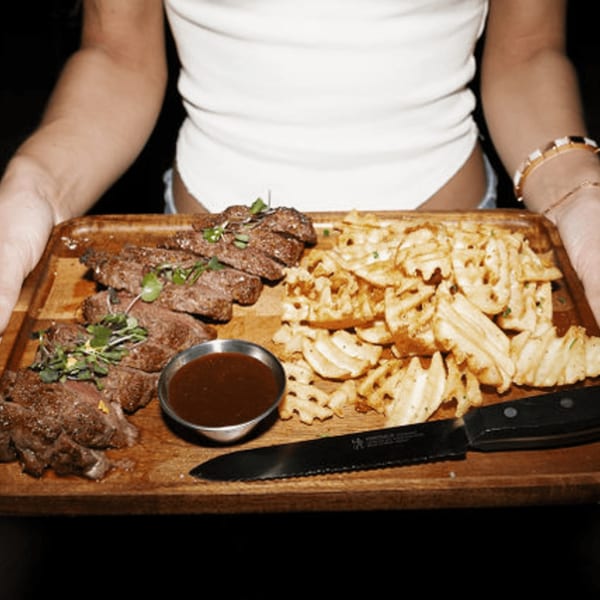 Hands holding a wooden tray with sliced steak, waffle fries, and a dipping sauce at Dodo Bird Kitchen.