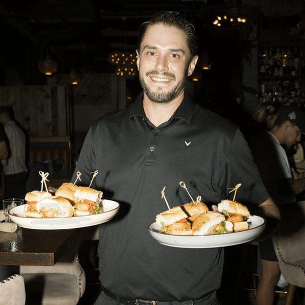 Smiling Dodo Bird Kitchen and Cocktails server holding two plates of mini sandwiches in a dimly lit bar.