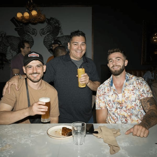 Three smiling men at Dodo Bird Kitchen and Cocktails, holding drinks at a bar.