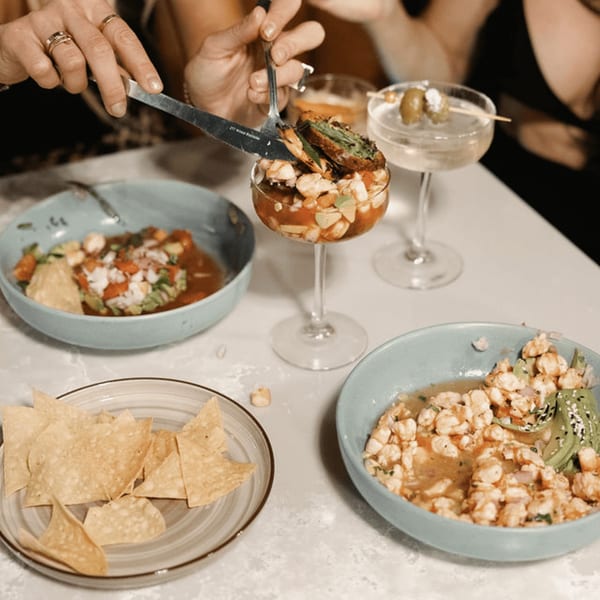 Hands serving ceviche from a glass, with other ceviche bowls, tortilla chips, and cocktails on a table.