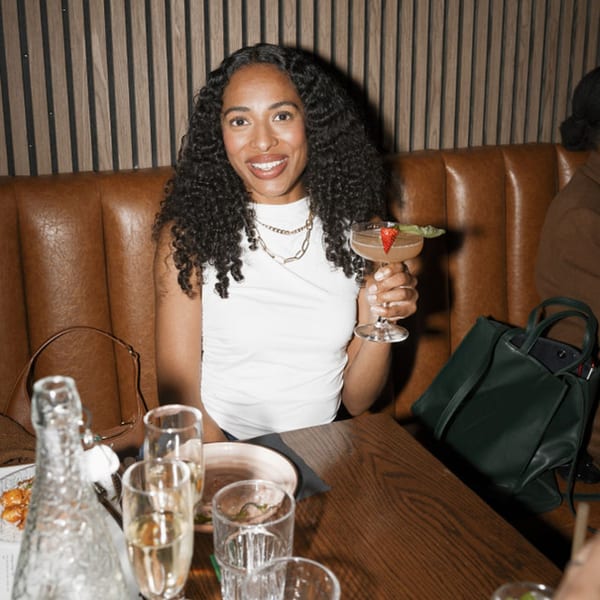 A smiling woman with curly hair holds a cocktail at a Dodo Bird Kitchen and Cocktails booth.