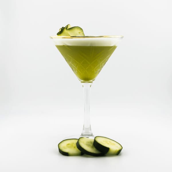 Green cocktail in a martini glass with cucumber garnish and slices on a white background.