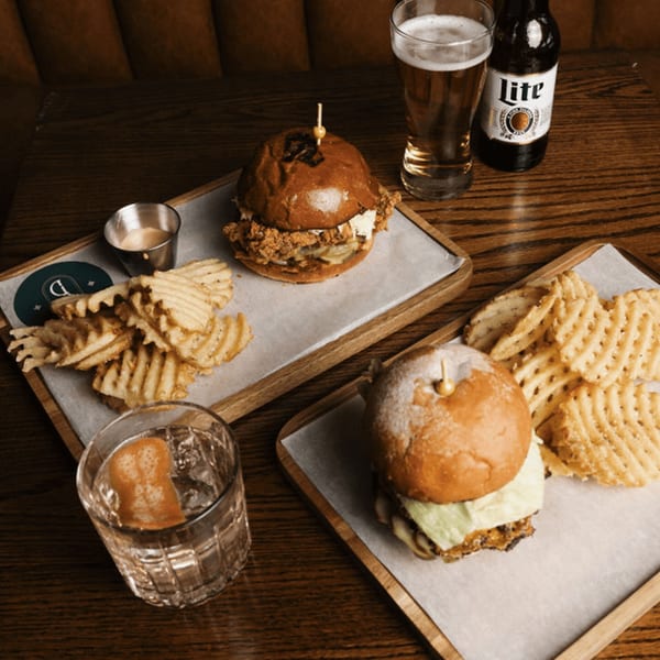 Two Dodo Bird Kitchen burgers with waffle fries, a beer, and a cocktail on a wooden table.