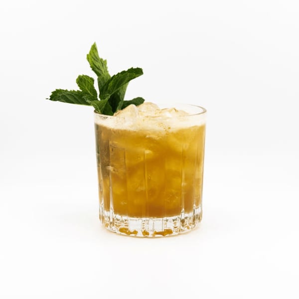 A Dodo Bird Kitchen and Cocktails cocktail, amber-colored with ice and a mint garnish, on a white background.