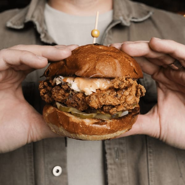Hands holding a Dodo Bird Kitchen fried chicken sandwich with pickles, slaw, and sauce.